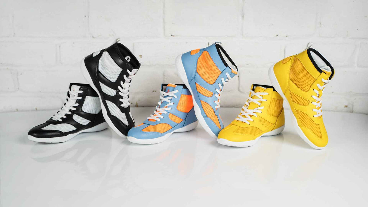 Four high-top sneakers arranged in a row against a white brick wall and white surface: a black-and-white pair, a light blue-and-orange pair, a yellow-and-white pair, and a bright yellow pair.